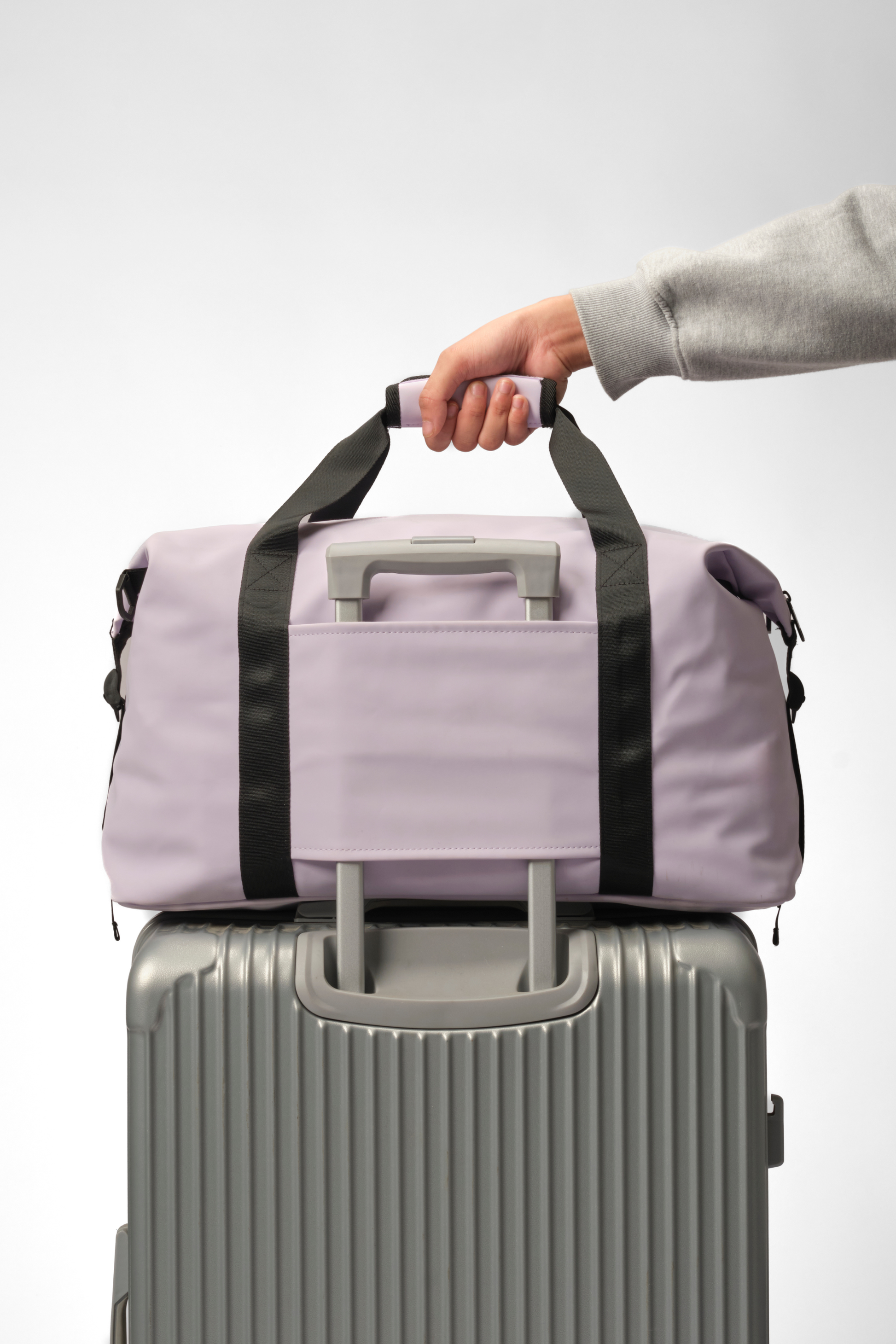 A lavender purple duffel bag with black handles and gray accents, shown resting on top of a silver rolling suitcase. A person's hand and sleeve are visible holding the bag's handle, set against a clean light gray studio background.