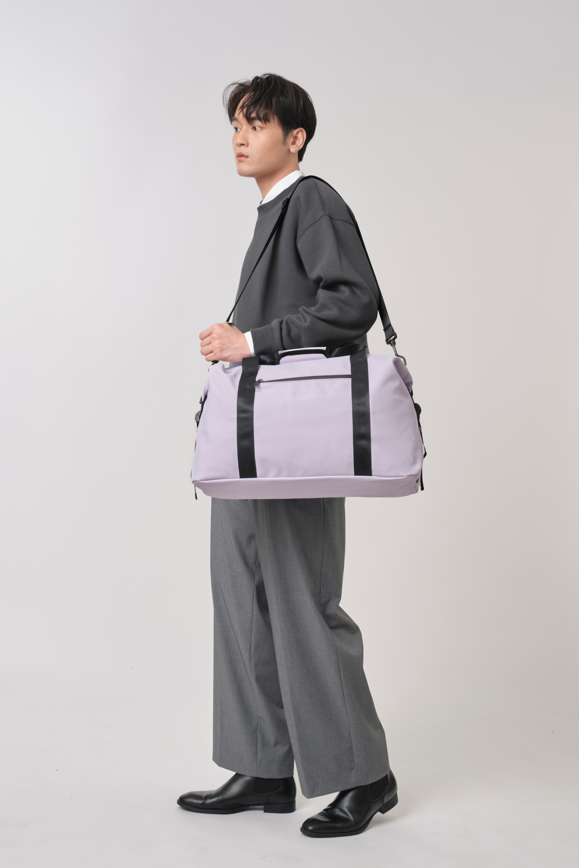 A man in a dark gray blazer, white collared shirt, and gray trousers carries a light purple duffel bag with black straps and trim against a light gray studio background.